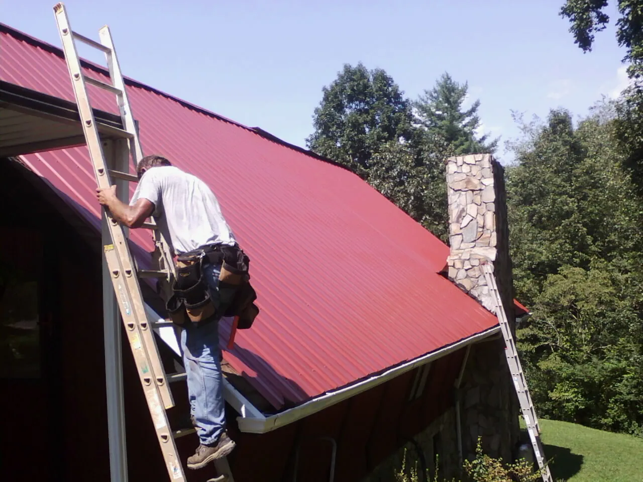 Roofer on a ladder beside a red metal roof and stone chimney in Brookeville